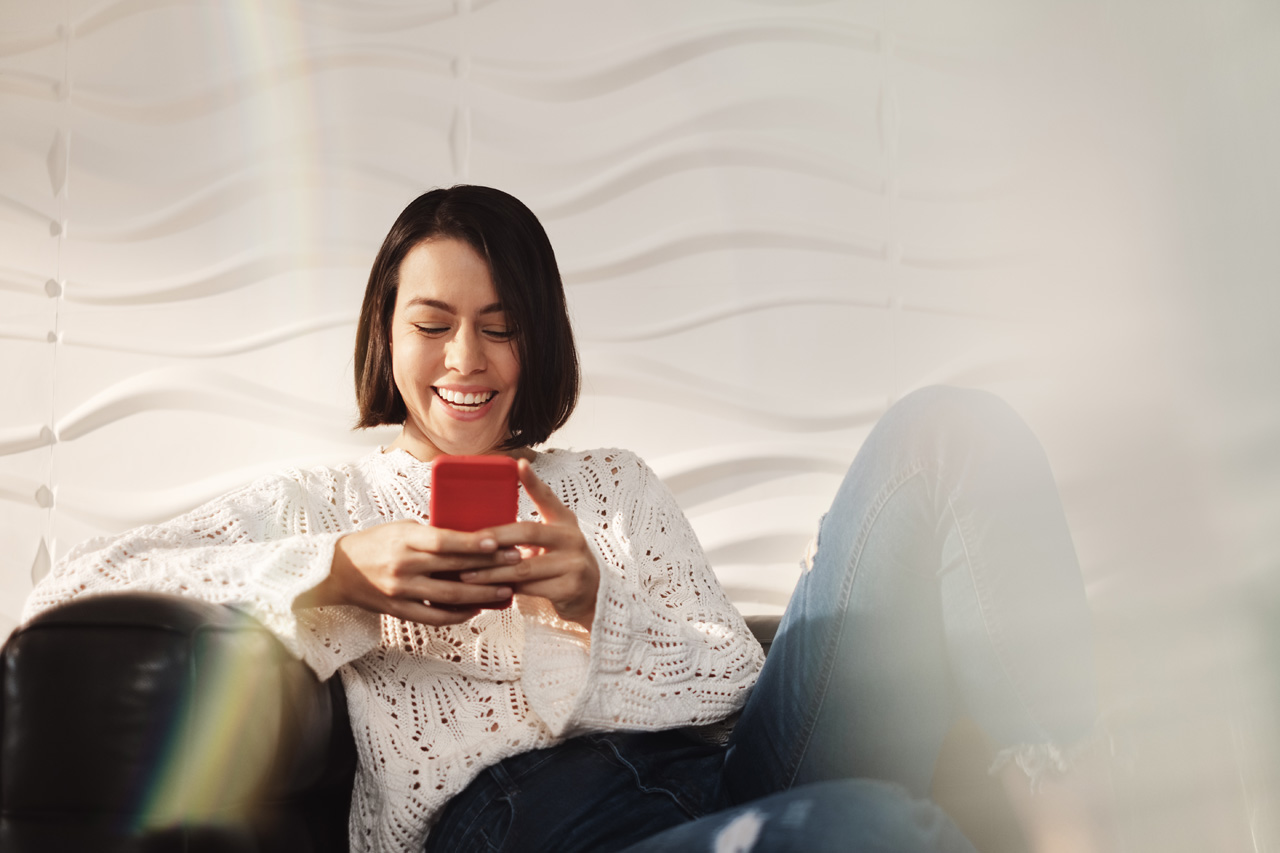 Real estate broker smiling while sitting on a couch and using her phone to access apps and tools.